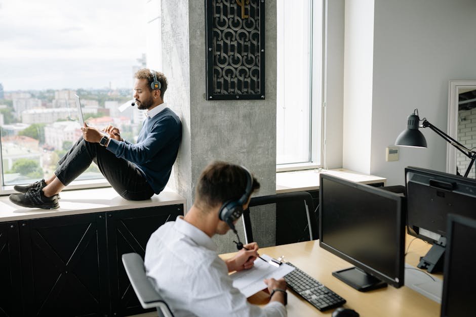Two call center agents working in a modern office, one seated at a computer and another using a tablet by the window.