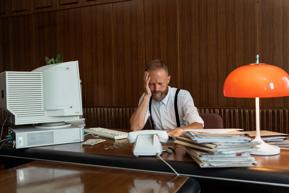 Bearded adult man feeling stressed at vintage office desk with old computer and paperwork.